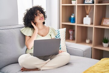 Canvas Print - Young brunette woman with curly hair using laptop sitting on the sofa at home smiling with hand over ear listening an hearing to rumor or gossip. deafness concept.