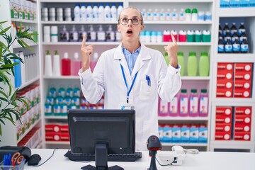 Poster - Young caucasian woman working at pharmacy drugstore amazed and surprised looking up and pointing with fingers and raised arms.