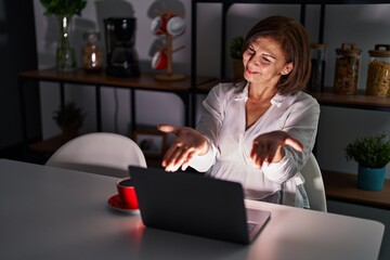 Poster - Middle age hispanic woman using laptop at home at night looking at the camera smiling with open arms for hug. cheerful expression embracing happiness.