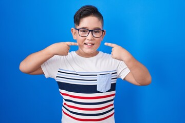 Sticker - Young hispanic kid standing over blue background smiling cheerful showing and pointing with fingers teeth and mouth. dental health concept.