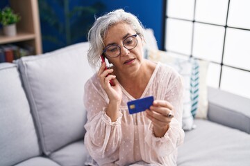Canvas Print - Middle age grey-haired woman talking on smartphone holding credit card sitting on sofa at home