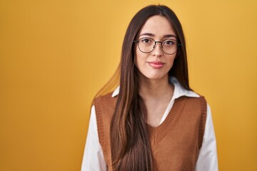 Poster - Young brunette woman standing over yellow background wearing glasses relaxed with serious expression on face. simple and natural looking at the camera.