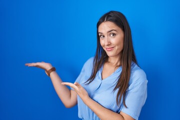 Poster - Young brunette woman standing over blue background inviting to enter smiling natural with open hand