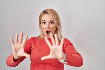 Wall Mural - Blonde woman standing over isolated background afraid and terrified with fear expression stop gesture with hands, shouting in shock. panic concept.