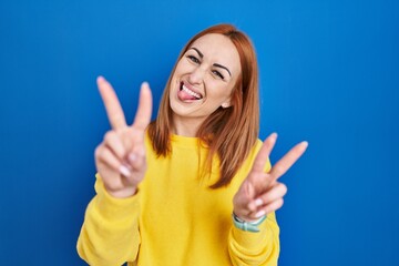 Poster - Young woman standing over blue background smiling with tongue out showing fingers of both hands doing victory sign. number two.