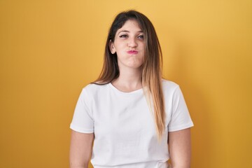 Wall Mural - Young brunette woman standing over yellow background puffing cheeks with funny face. mouth inflated with air, crazy expression.