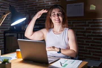 Wall Mural - Brunette woman working at the office at night gesturing with hands showing big and large size sign, measure symbol. smiling looking at the camera. measuring concept.