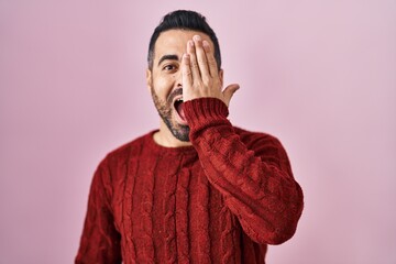 Wall Mural - Young hispanic man with beard wearing casual sweater over pink background covering one eye with hand, confident smile on face and surprise emotion.