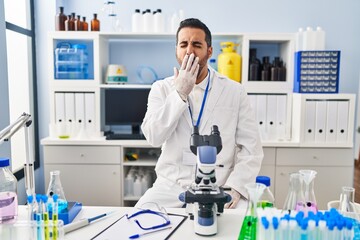 Canvas Print - Young hispanic man with beard working at scientist laboratory bored yawning tired covering mouth with hand. restless and sleepiness.