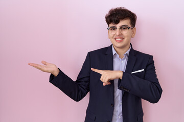 Wall Mural - Young non binary man with beard wearing suit and tie amazed and smiling to the camera while presenting with hand and pointing with finger.