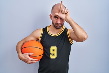 Poster - Young bald man with beard wearing basketball uniform holding ball making fun of people with fingers on forehead doing loser gesture mocking and insulting.