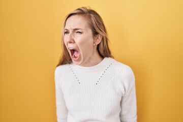 Canvas Print - Young caucasian woman wearing white sweater over yellow background angry and mad screaming frustrated and furious, shouting with anger. rage and aggressive concept.