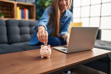 Wall Mural - Young blonde woman inserting coin on piggy bank sitting on sofa at home