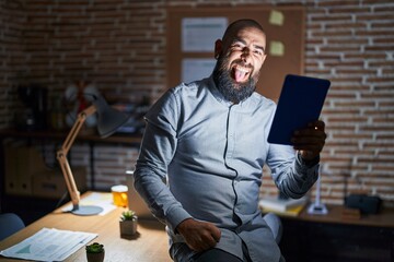 Canvas Print - Young hispanic man with beard and tattoos working at the office at night sticking tongue out happy with funny expression. emotion concept.