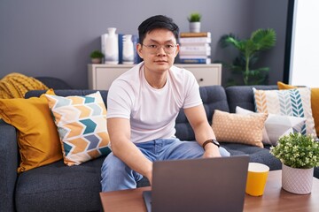 Sticker - Young asian man using laptop at home sitting on the sofa relaxed with serious expression on face. simple and natural looking at the camera.