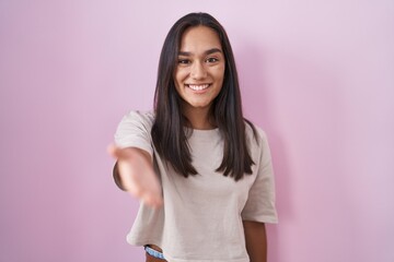 Wall Mural - Young hispanic woman standing over pink background smiling cheerful offering palm hand giving assistance and acceptance.