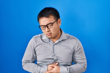 Canvas Print - Young chinese man standing over blue background with hand on stomach because nausea, painful disease feeling unwell. ache concept.