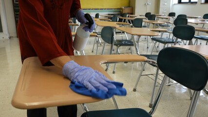 Pretty woman teacher with spray bottle, gloves, rag, wiping, cleaning and disinfecting school desks.