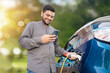 © GAJENDRRA BHATI  - Happy Indian man attaching power cable into socket to charge his modern electric car at outdoor charging station while using smart phone. Electric vehicle Recharging battery charging port.
