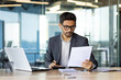 © Liubomir - Serious concentrated hispanic businessman financier on paperwork inside office, man in business suit at workplace reviewing and reading papers, contracts and accounts reports