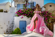 Pretty woman in pink long romantic dress is posing in Oia, Santorini Island. Female model in amazing long dress. Couture.