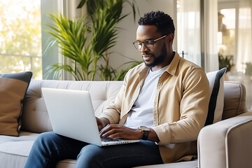 A young African American man works as a neural network tutor. A man sits on the couch with a laptop on his lap and teaches online.