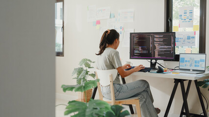 Side view of professional young Asia girl IT development programmer typing on keyboard coding programming fixing data code on computer screen and laptop on table in workroom at house office.