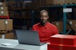 African man in red shirt in delivery service warehouse, looking at camera, sitting at office desk with laptop. Using a laptop while working. Generative ai.