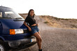 A Latina girl with braided hair in casual clothes is leaning on a camper van while looking at the camera happily. Concept of African women traveling in summer to the mountains. Copyspace.