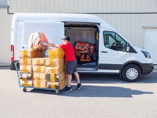 Loading goods into minivan. Man is loading boxes into car. Transport company specialist. Guy is loading boxes into minivan. Delivery man near cargo trolley. Logistician in industrial zone