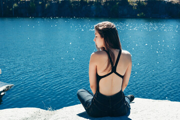 A beautiful woman sits on a rock cliff near the canyon lake on a background of shining water from the sun in a warm sunny day.