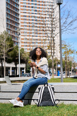 Wall Mural - Young happy African American woman sitting in city park using mobile phone shopping online or texting messages. Smiling female student holding cellphone chatting in sunny campus area, vertical.