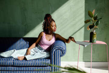 Beautiful African woman wearing casual clothes, reaching for the magazine while sitting on the sofa in a living room with sunlight. Portrait of a relaxed female resting at home. Copy space for text.