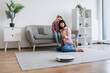 © sofiko14 - Caring woman cuddling preteen girl on sofa while sitting on floor with switched on robot vacuum cleaner. Mother-daughter duo sharing valuable moments while smart device removing dust from carpet.