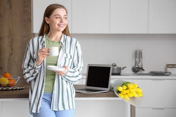 Sticker - Beautiful young woman drinking coffee near table with modern laptop and bouquet of yellow tulip flowers