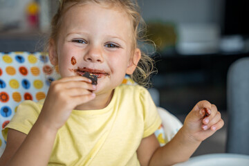 Wall Mural - Little girl with blond hair eating homemade chocolate with dirty mouth and hands in home kitchen