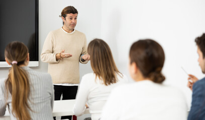 Male business coach explaining topic while standing against group of people sitting at desks in front of him