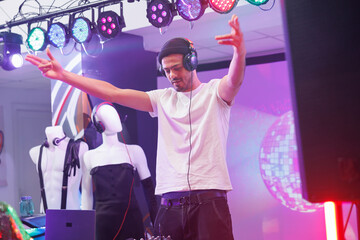 Young dj in headphones performing on stage and raising hands while mixing electronic music in club. Man musician wearing earphones playing in nightclub at discotheque party