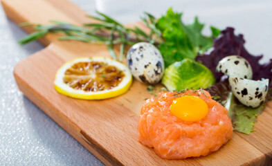 Tartare of fresh salmon on wooden board. Recipe: finely chop 200 gr of fish, season with salt, black pepper, olive oil, lemon juice, garnish with yolk of quail egg, greens and balsamic vinegar..