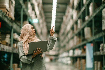 Wall Mural - Portrait of Asian muslim female headscarf hijab holding computer laptop checklist product shelves warehouse. woman worker inspecting goods industry. Logistic concept.