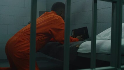 Wall Mural - African American prisoner in orange uniform kneels near the bed, prays to God in prison cell with Bible. Male criminal serves imprisonment term in jail or detention center. Concept of faith in God.