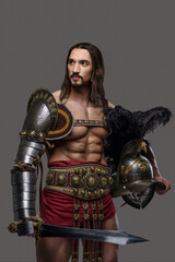 Muscular gladiator in graceful lightweight armor strikes a pose while holding a gladius and helmet, on a grey background