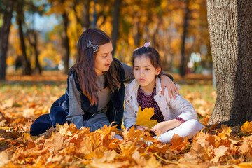 Wall Mural - two girls are sitting in a glade of yellow maple leaves in an autumn city park, children are playing and enjoying, picking leaves near a tree, beautiful nature, bright sunny day