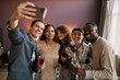 © Seventyfour - Happy young man with glass of red wine taking selfie with group of intercultural friends looking at camera while enjoying home party