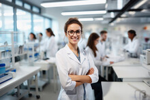 Beautiful Young Woman Scientist Wearing White Coat And Glasses In Modern Medical Science Laboratory With Team Of Specialists On Background. 