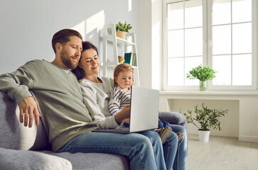 Happy young family with little child girl sitting on the sofa using modern laptop together. Smiling parents resting on couch enjoying weekend watching funny cartoon online or talking on video call.