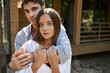 © LIGHTFIELD STUDIOS - Brunette man hugging girlfriend and looking at camera near blurred vacation house outdoors