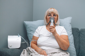 Wall Mural - Sick upset caucasian senior woman with an inhaler. Unhealthy female doing inhalation at home, she use nebulizer and inhaler for the treatment, sitting on the couch at home, need treatment. 