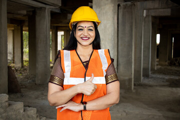 Portrait of confident Indian female civil engineer wearing helmet and vest standing at construction site do thumbs up with hand.