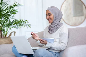 Arab young female student studying online at home via laptop. Sitting on the sofa in the lotus position, writing in a notebook, communicating via video call.